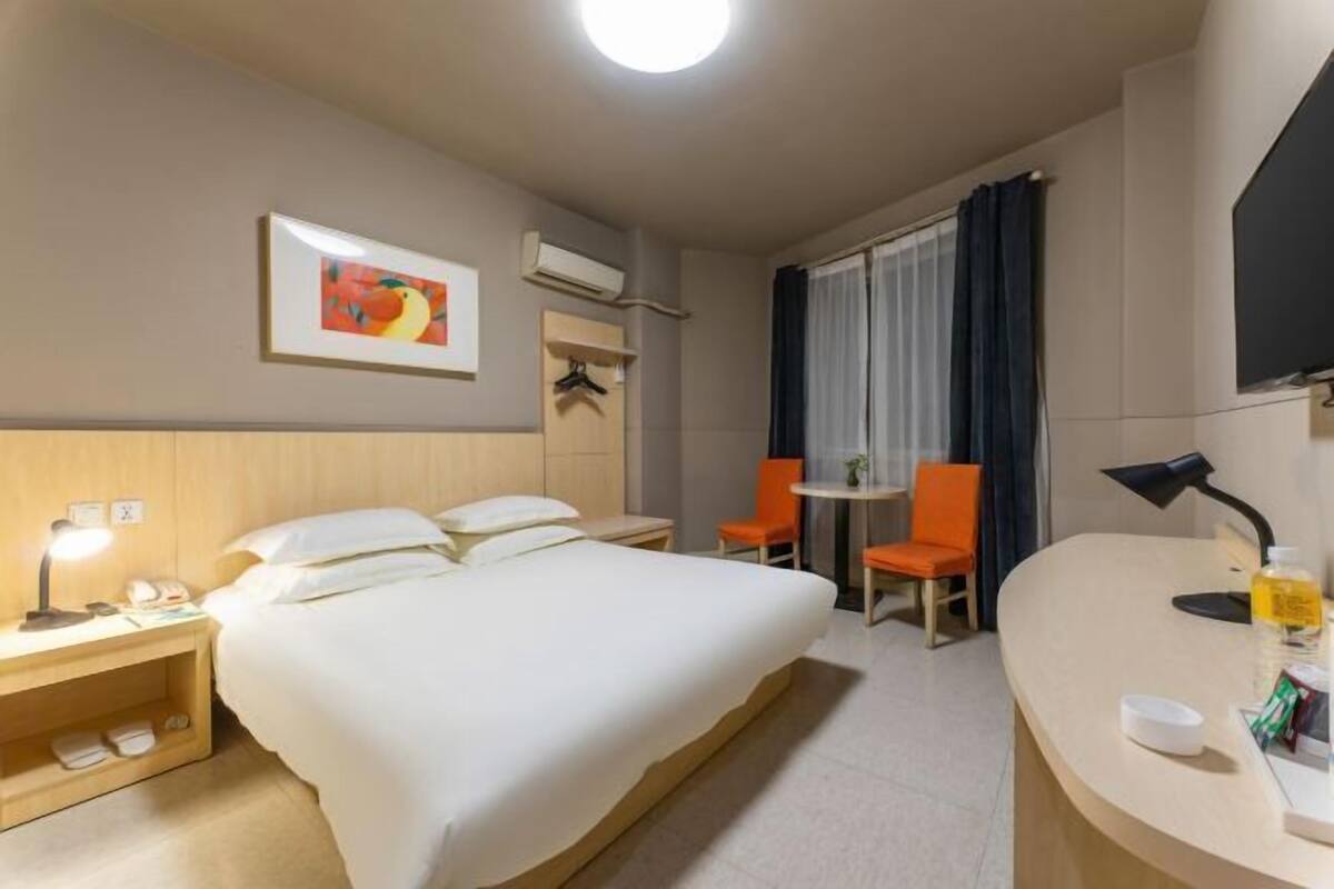 Una habitación de hotel con cama, mesita de noche, lámpara, silla, escritorio y un cuadro en la pared.