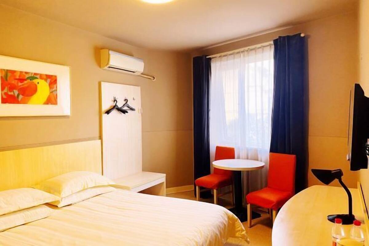 Habitación de hotel con dos camas, una mesita con sillas, un perchero y un ventanal con cortinas.