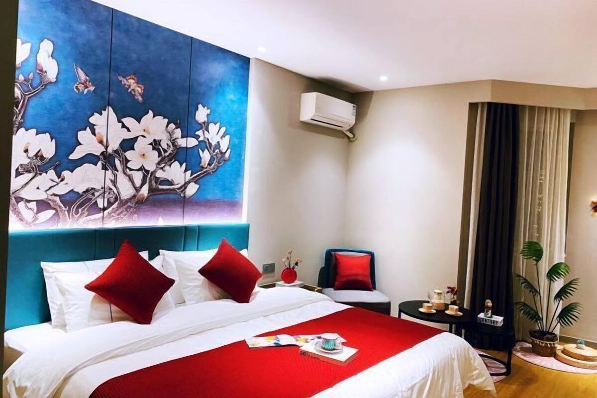 Habitación de hotel con una cama grande, ropa de cama roja y blanca, un cuadro de flores en la pared, una mesita con un florero y una planta en maceta.