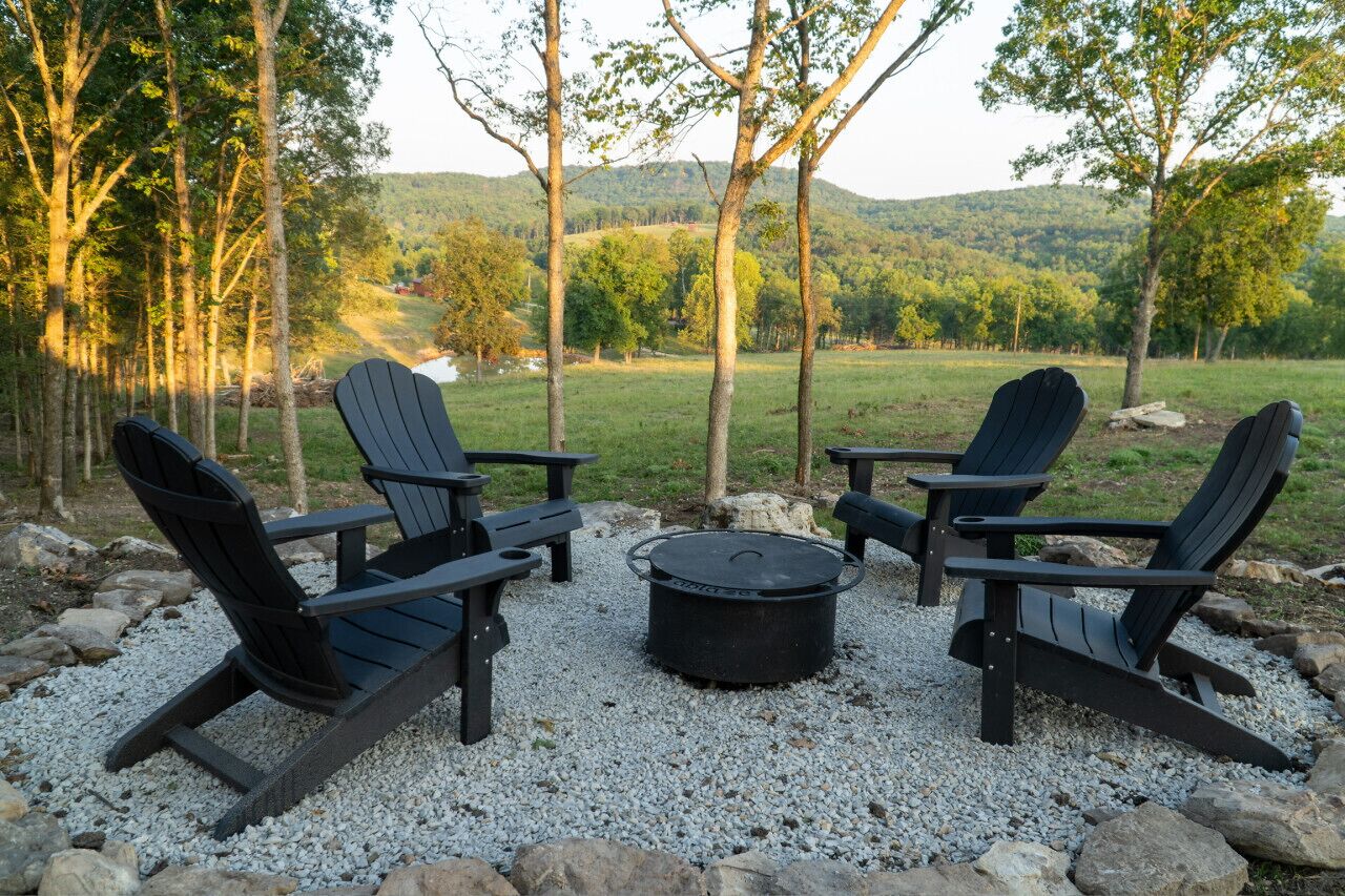 A seating area with Adirondack chairs and a fire pit surrounded by gravel, overlooking a forested landscape.