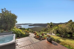 A wooden deck with a hot tub, surrounded by lush greenery and a view of the ocean. - Sunrise Lookout - Paihia Holiday Home (Haruru)