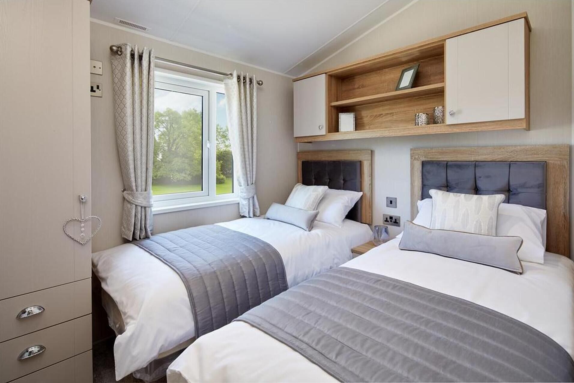 A bedroom with two beds, a wardrobe, a window with curtains, and a shelf above the beds.