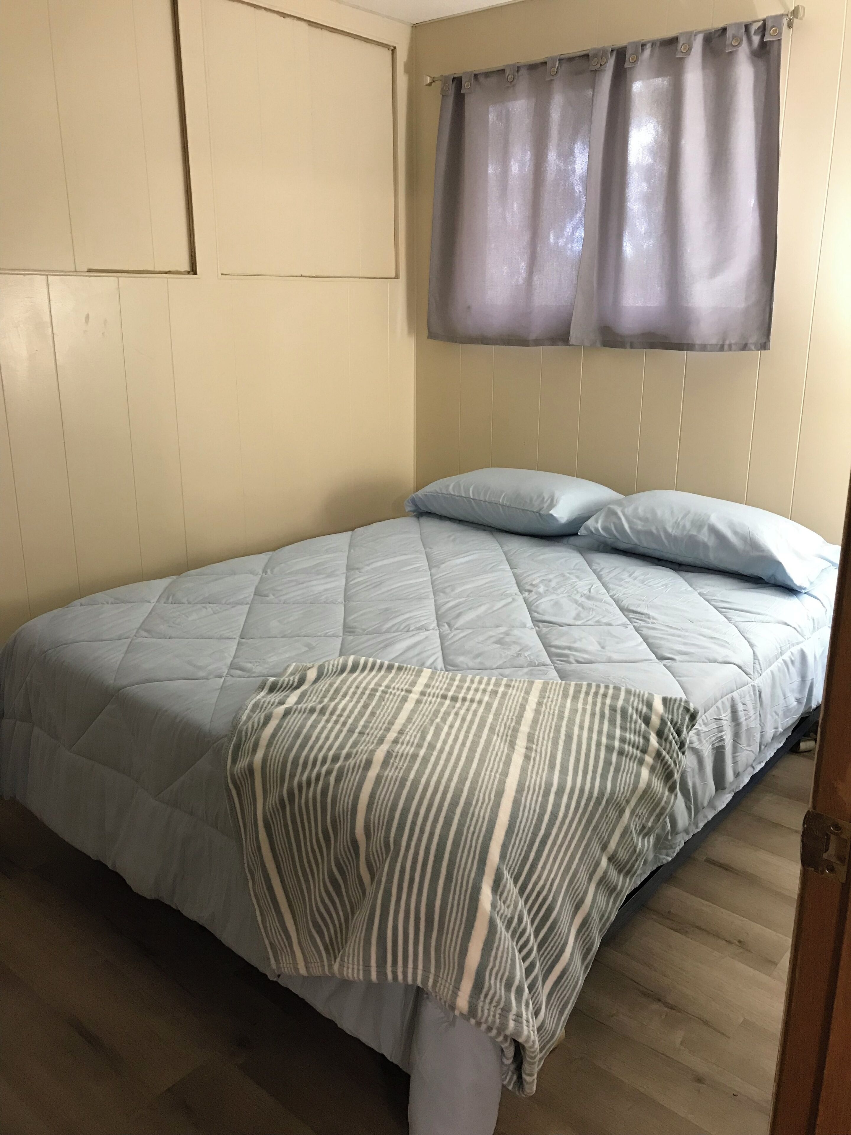 A single bed with a quilted blue bedspread and two pillows, a striped throw blanket, and a window with sheer curtains.
