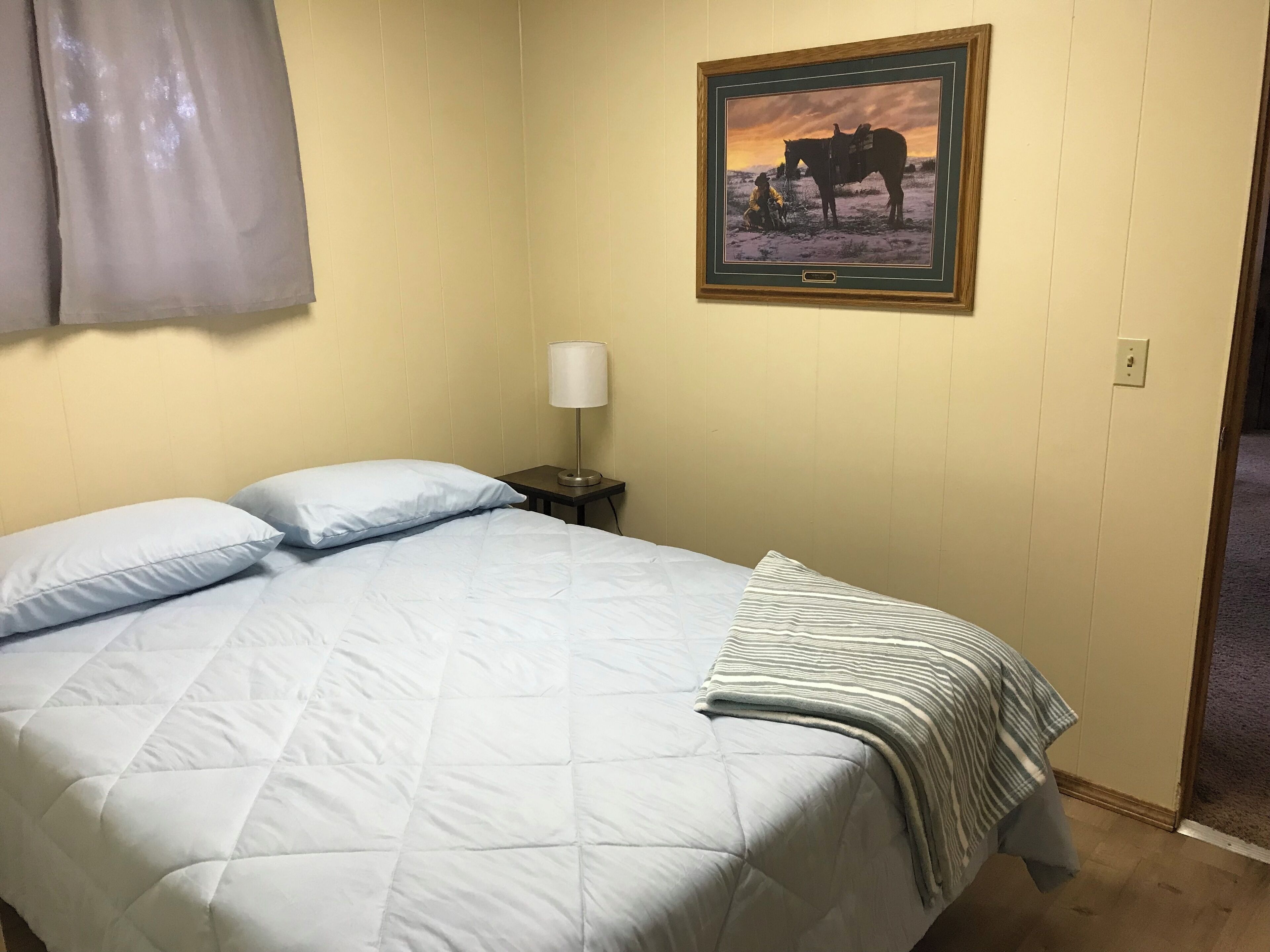 A neatly made bed with a quilted cover and pillows, a bedside lamp, and a framed picture of a horse on the wall.