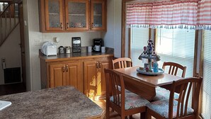 A kitchen with wooden cabinets, a dining table with chairs, a clock, and a window with blinds. - 1 bedroom, 2 loft Park Model in campground in Marquette, Iowa.  (Marquette)