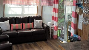 A cozy living room with a dark leather sofa, a ceiling fan, and a window with striped curtains. - 1 bedroom, 2 loft Park Model in campground in Marquette, Iowa.  (Marquette)