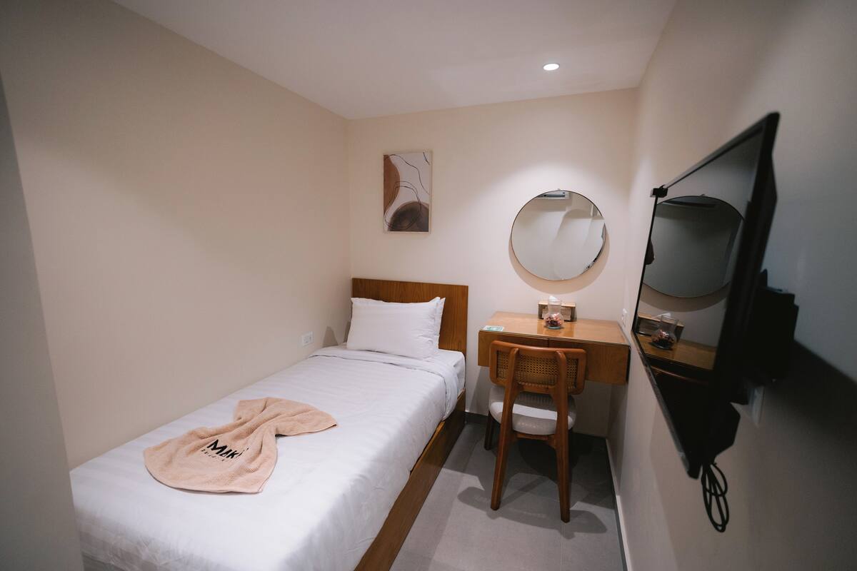 A small, neatly arranged bedroom with a single bed, a wooden desk, a chair, a round mirror, and a television mounted on the wall.