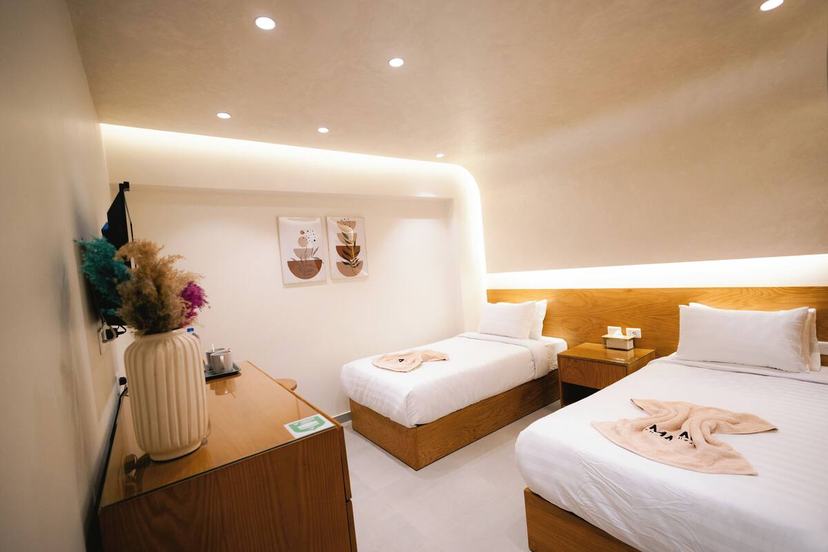 A modern hotel room with two beds, a wooden nightstand, a desk with a vase, and wall art.