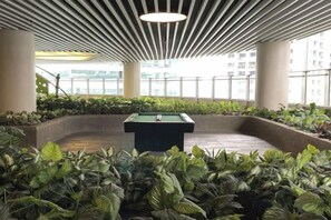 Een pooltafel in een ronde plantenbak, omgeven door weelderige begroeiing, met een modern plafondontwerp en grote ramen op de achtergrond.