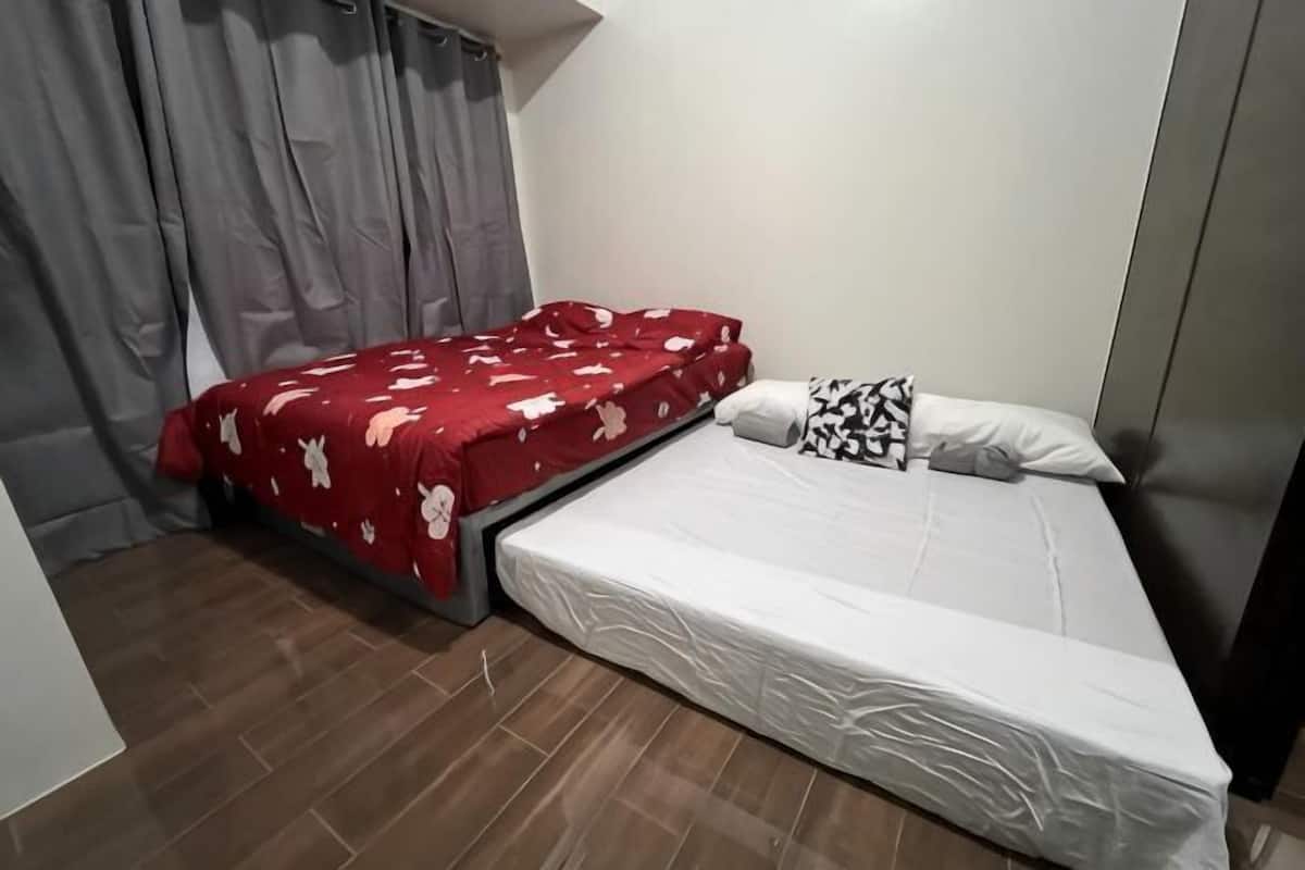 Una habitación con una cama, una colcha roja, un colchón blanco y una cortina gris.