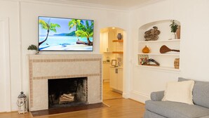 A living room with a fireplace, a television mounted above it, a grey sofa, and a kitchen area in the background. - Intercoastal escape, 1mi beach, pool, downtown, private yard (West Palm Beach)