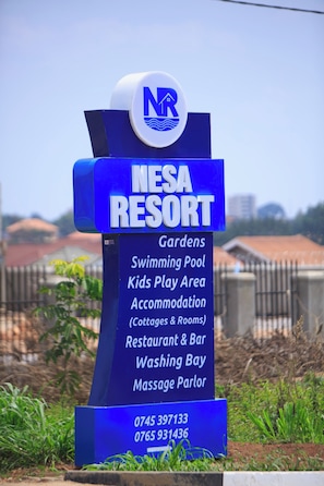 Un letrero para Nesa Resort que enumera servicios como jardines, piscina, área de juegos para niños, alojamiento, restaurante y bar, y salón de masajes.