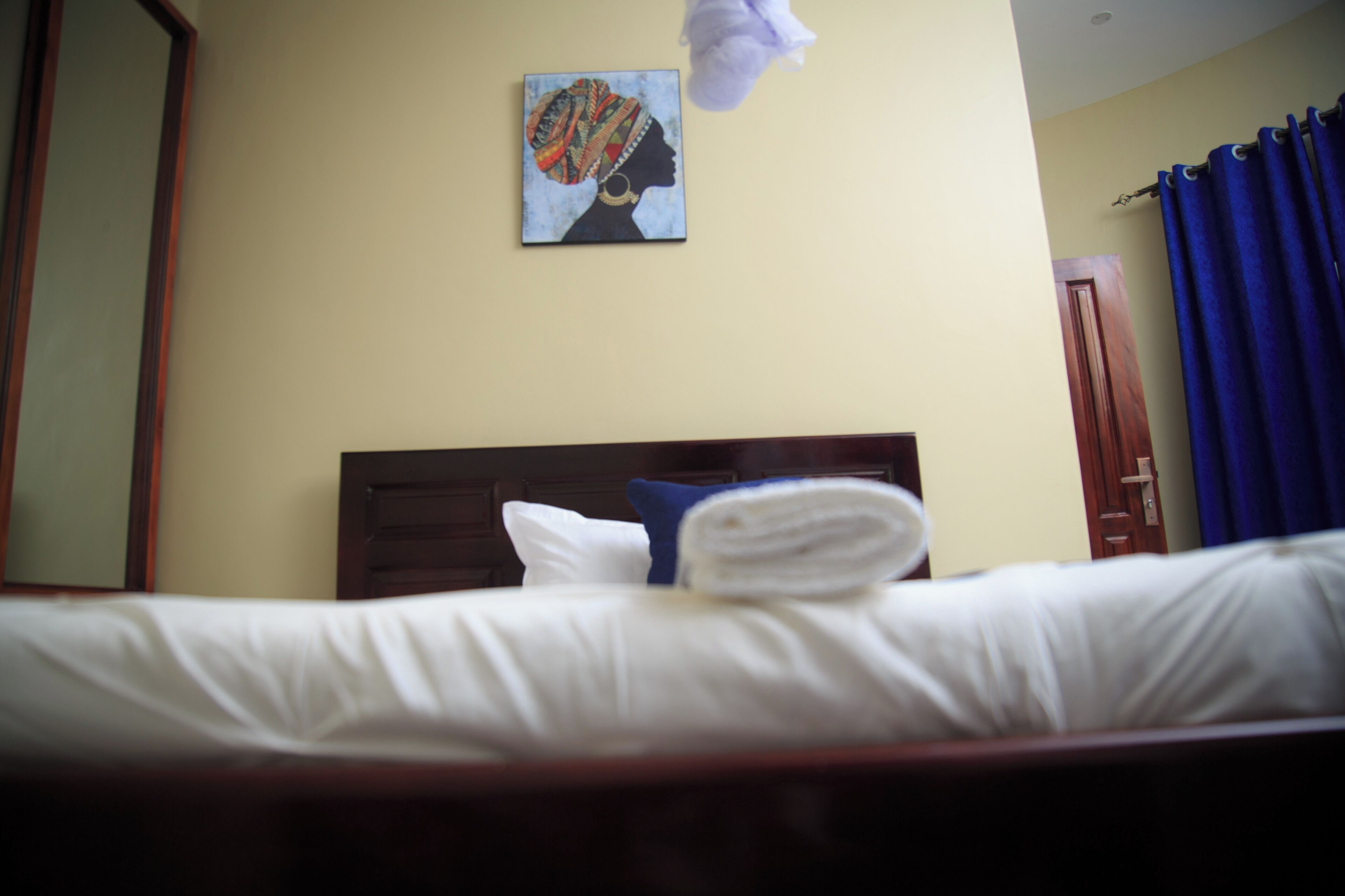 A hotel room with a neatly made bed, a headboard, a painting on the wall, and a blue curtain.