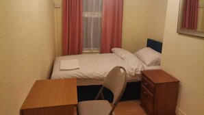 A small, single-bed room with a wooden desk and chair, a bedside table, and a window with curtains. - Single bedroom near London Seven Kings Train Station (Ilford)