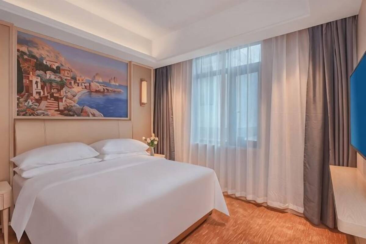 A hotel room with a large bed, a painting on the wall, a window with curtains, and a nightstand.