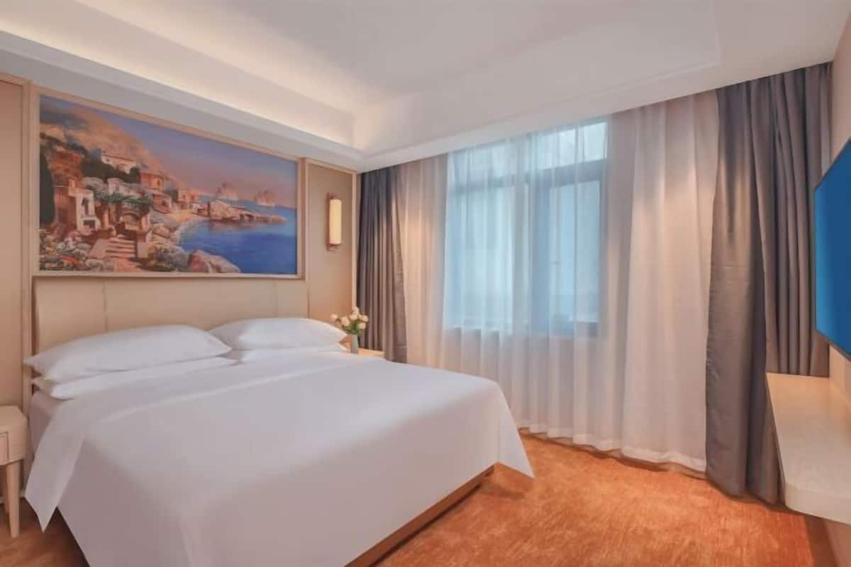 A hotel room with a large bed, a painting of a coastal scene, a window with sheer curtains, and a nightstand with a vase of flowers.