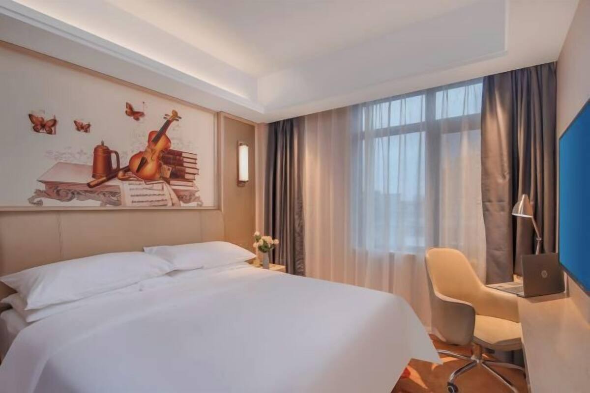 A hotel room with a bed, a desk with a laptop, a chair, a large window with curtains, and a wall mural.