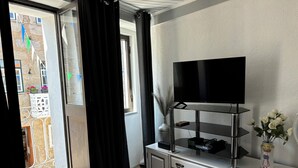 Une pièce avec un téléviseur posé sur une étagère en verre, un meuble blanc avec des tiroirs, un vase avec des fleurs et une vue sur un balcon avec une porte et des rideaux.