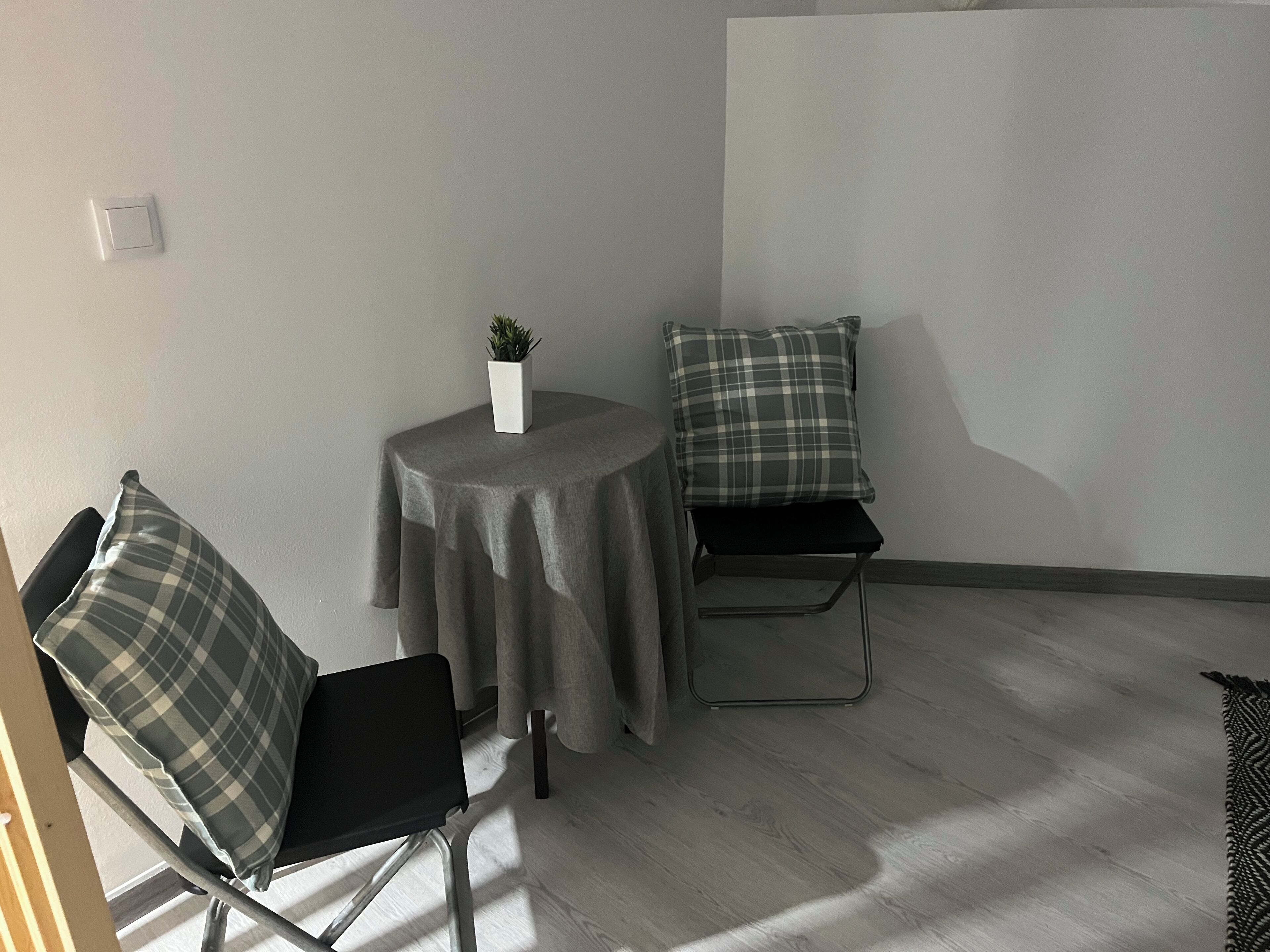 Une petite pièce, bien rangée, avec une table ronde, deux chaises, une plante en pot et un coussin à carreaux.