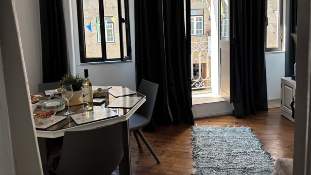 Un coin repas avec une table dressée pour deux personnes, un ordinateur portable et une bouteille de vin. Les fenêtres sont ornées de rideaux noirs, et on peut apercevoir le balcon extérieur.