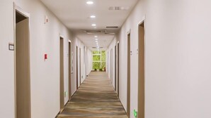 A long, narrow hotel corridor with doors on both sides, a patterned carpet, and a view of greenery through a window at the end.
