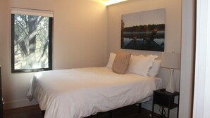 Une chambre avec un grand lit, une table de chevet, une lampe, une fenêtre donnant sur des arbres et un tableau au mur.