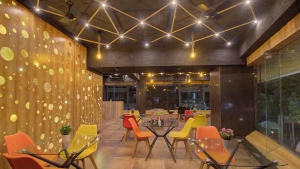 A modern interior with a central glass table surrounded by colorful chairs, a wooden wall with circular patterns, and a ceiling with exposed beams and hanging lights. - Greenstones Boutique Hotel (Nashik)