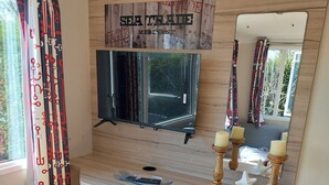 A cozy room with a wooden wall, a mounted TV, a mirror, and a shelf with books and candles.