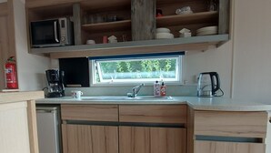 A kitchen with wooden cabinets, a microwave, a coffee maker, a kettle, and a window with a view of greenery.