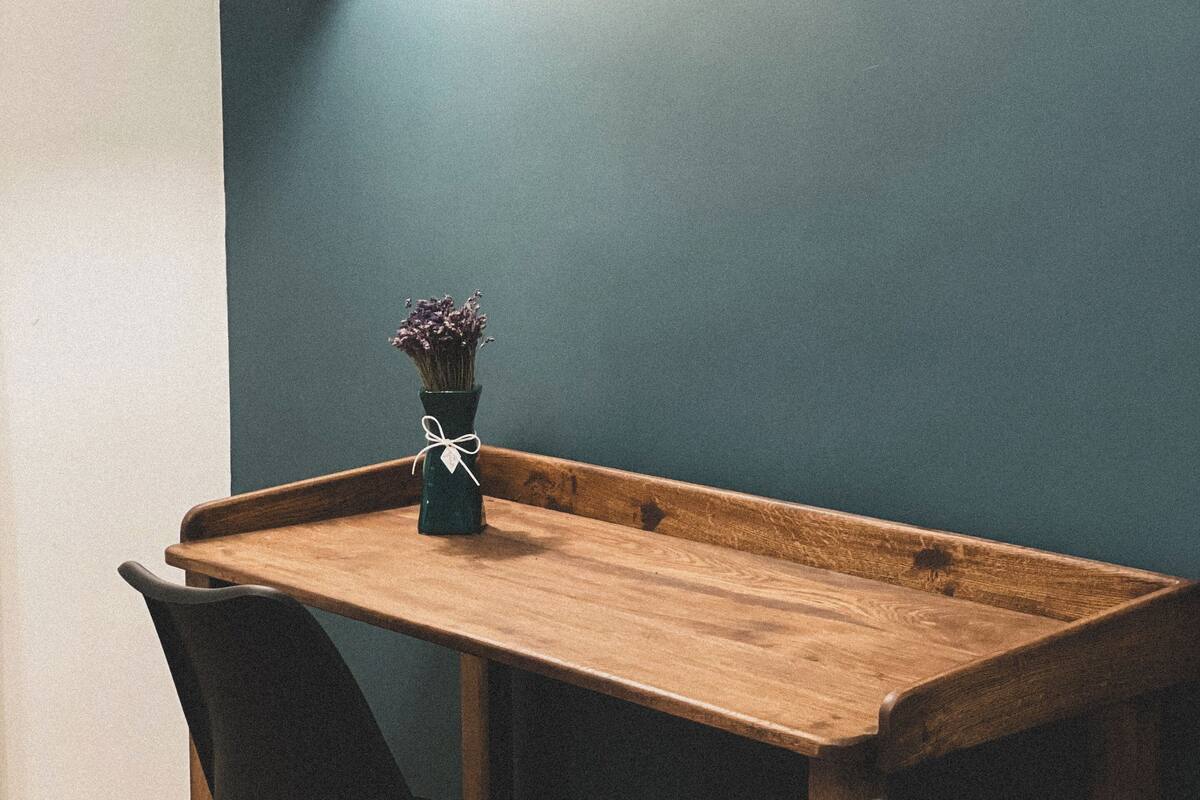 Une table en bois avec deux chaises, un vase de fleurs, une lampe fixée au mur et un petit cadre photo sur un mur de couleur turquoise.