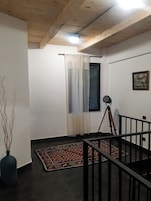Una habitación con techo de madera, una ventana con cortinas, un trípode, un cuadro en la pared y una alfombra estampada en el suelo.