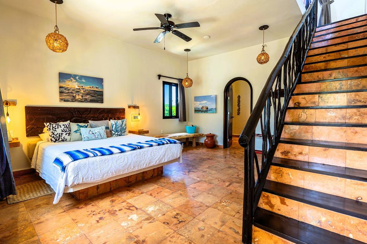 A bedroom with a bed, a ceiling fan, a window, and a staircase.