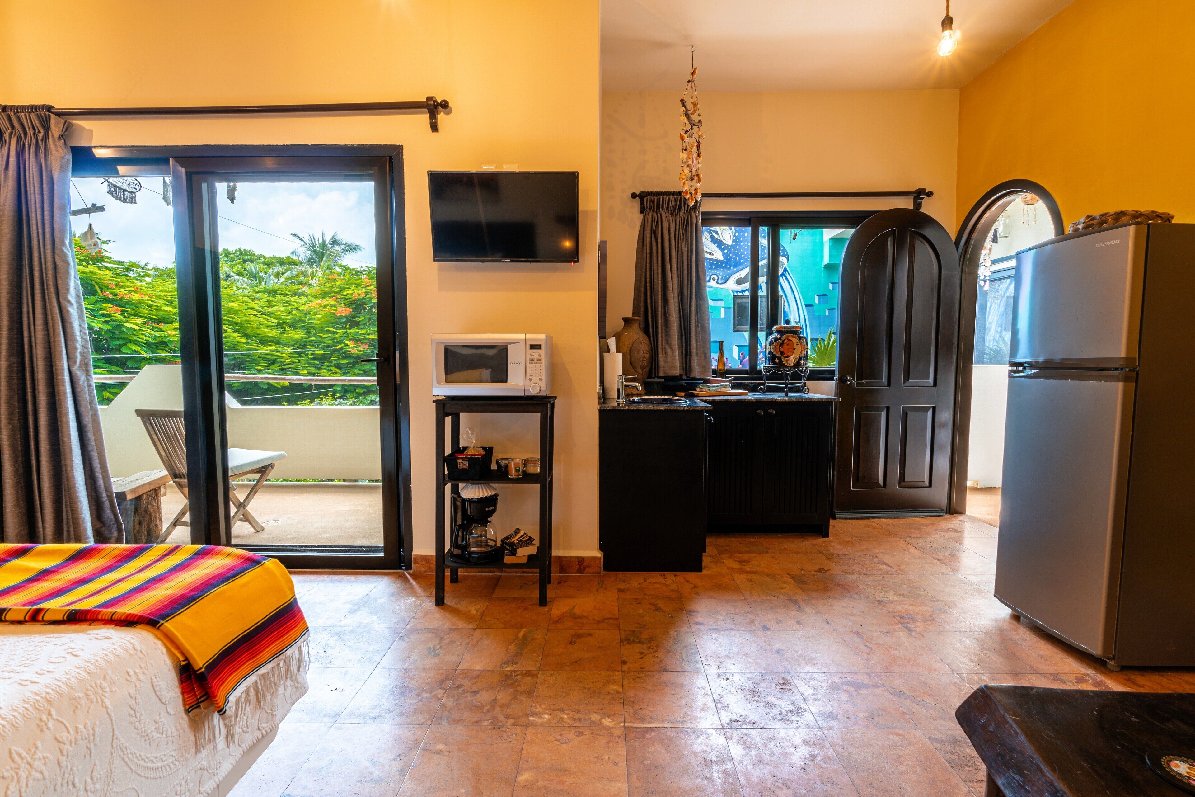 A hotel room with a bed, a small kitchenette, a TV, and a balcony with a view of greenery.