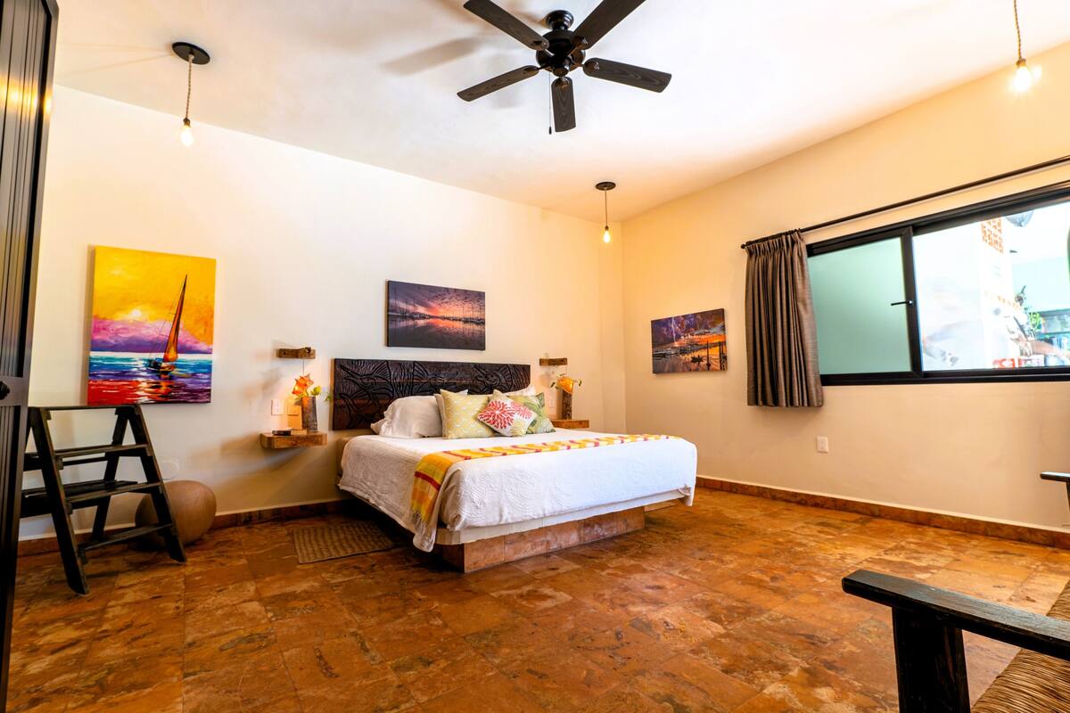 A bedroom with a bed, a ceiling fan, artwork on the walls, a window with curtains, and a wooden floor.