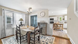 A dining area with a wooden table, chairs, and a rug. There is a clock on the wall and a kitchen in the background. - Charming 4-bed 2- Bath Home Close Proximity to Princeton Area and Sesame Place (Lawrence Township)