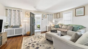 A living room with a sofa, a coffee table, a TV, and a fireplace. There are curtains, a plant, and a picture frame on the wall. - Charming 4-bed 2- Bath Home Close Proximity to Princeton Area and Sesame Place (Lawrence Township)