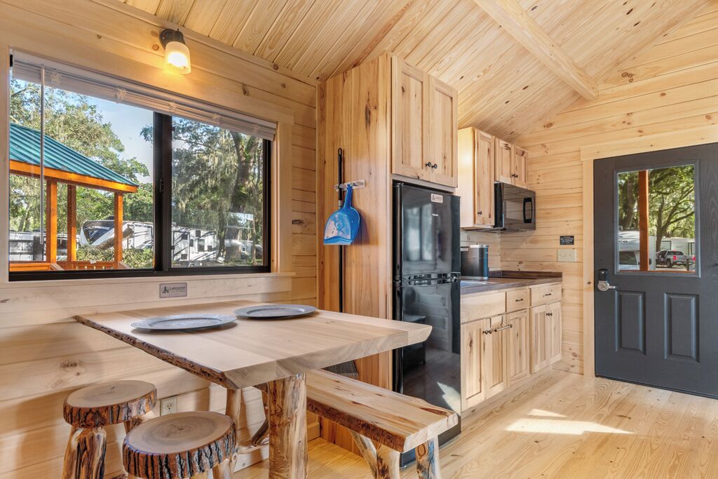 A wooden cabin kitchen with a large window, a dining table with stools, and a refrigerator.