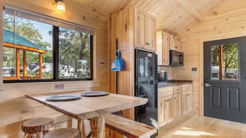 A wooden cabin kitchen with a large window, a dining table with stools, and a refrigerator.