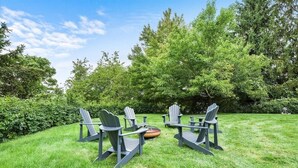 A grassy outdoor area with four Adirondack chairs arranged in a semi-circle, a fire pit in the center, and dense trees in the background. - Modern and Spacious House (Pittsburgh)