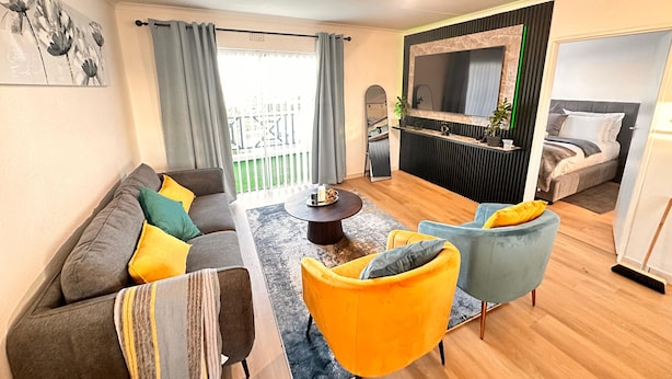 Ein modernes Wohnzimmer mit einer Couch, Sesseln, einem Couchtisch und einem Fernsehbereich, im Hintergrund ein Bett.