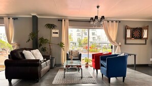 Ein modernes Wohnzimmer mit einer Ledersofa, einem blauen Sessel und einem Glastisch. Ein großes Fenster mit Vorhängen, eine Pflanze und ein Dartspiel an der Wand.