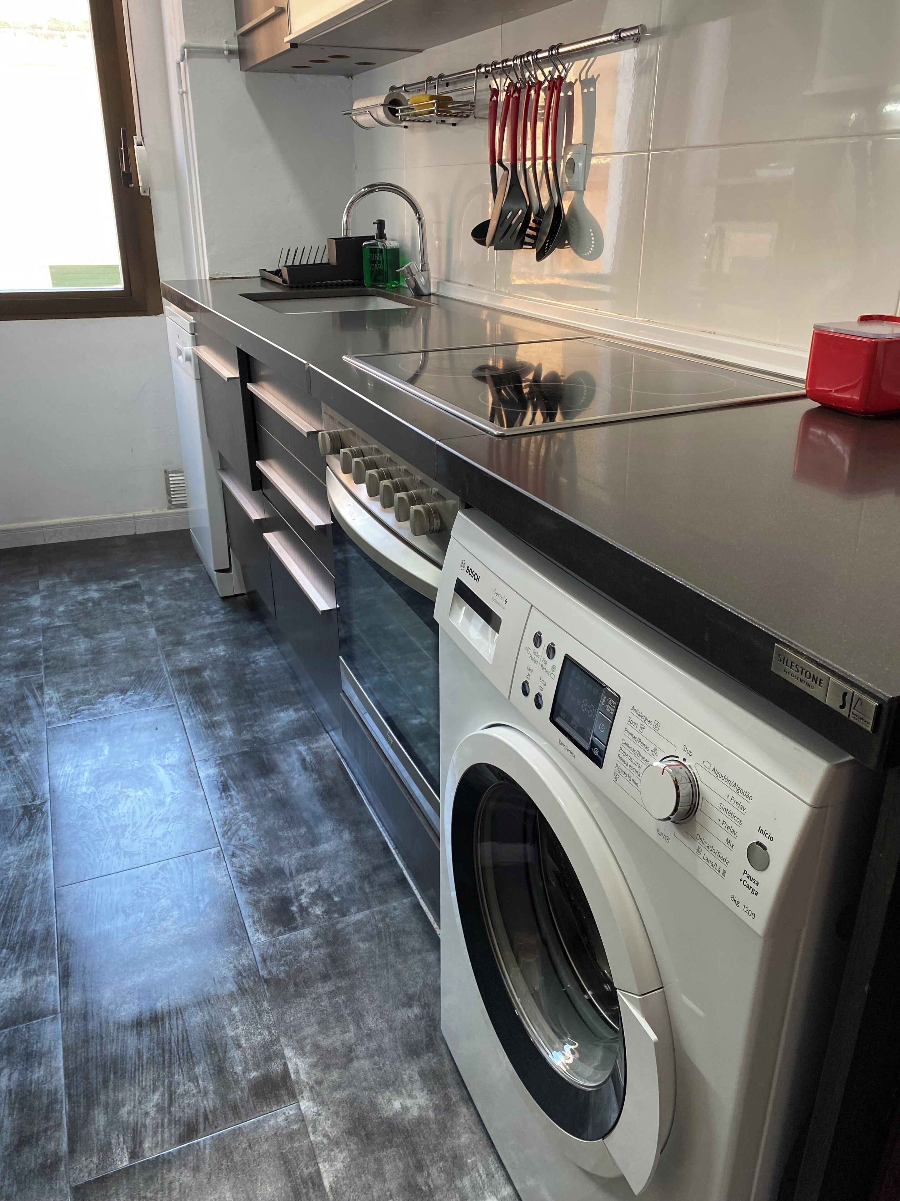 A modern kitchen with a Bosch washing machine, stainless steel countertops, and a window providing natural light.