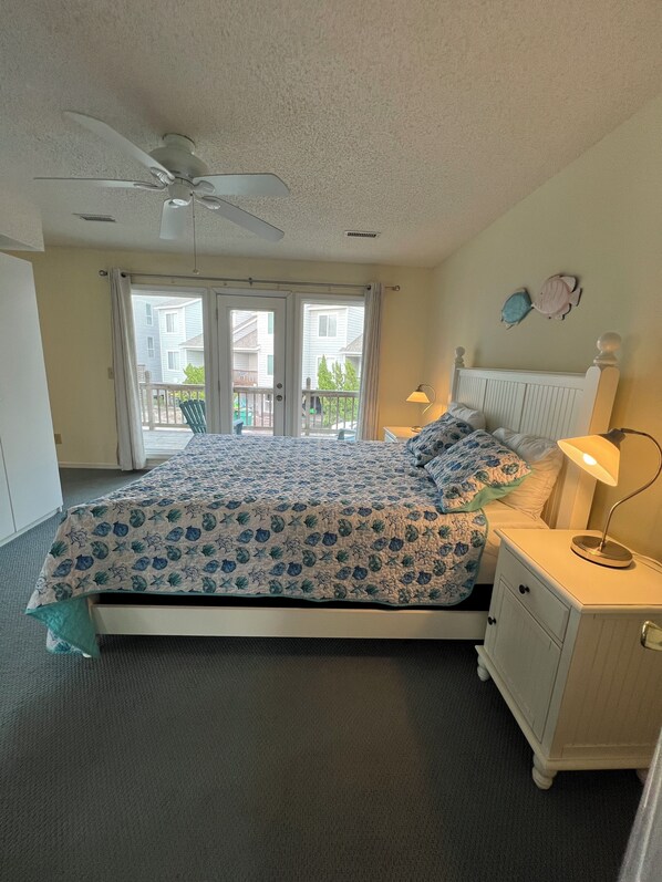 A bedroom with a bed, a nightstand, a lamp, a ceiling fan, and a view of the outside through a sliding door.