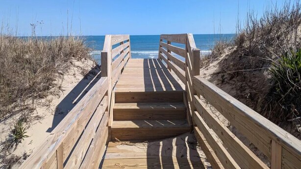 Ein hölzerner Steg führt zu einem Strand bei blauem Himmel.