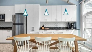 Eine moderne Küche mit weißen Schränken, einem Edelstahl-Kühlschrank, einem Holzespeisetisch mit weißen Stühlen und blauen Pendelleuchten.