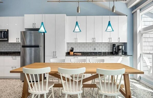A modern kitchen with white cabinets, a stainless steel refrigerator, a wooden dining table with white chairs, and blue pendant lights.
