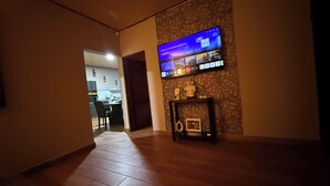 A modern living room with a flat-screen TV mounted on a stone wall, a small console table with decorative items, and a dining area visible through an open doorway.