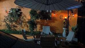 An outdoor seating area with a large umbrella, chairs, tables, potted plants, and wall-mounted lights.