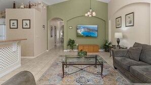 A spacious living area with a high ceiling, a large flat-screen TV mounted on the wall, a sofa, a coffee table, and a dining area with a kitchen counter. - Wookiee's Hideout - 4 bed private pool home in Cumbrian Lakes - 18 mins to WDW! (Kissimmee)