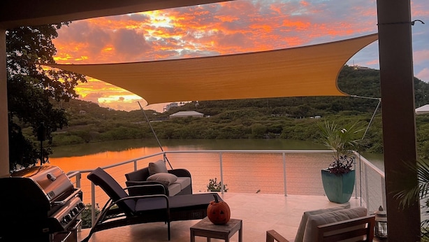A balcony with a view of a lake and sunset. There are lounge chairs, a table, and a grill.